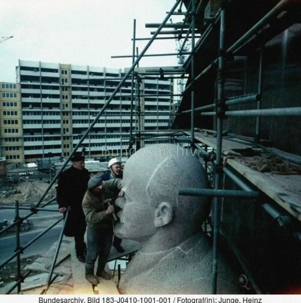 Photo of three workers finishing sculpting a Lenin's statue in a construction place, in front of an old huge soviet building. The image has a watermark of the Bundesarchiv (the German National Archive) and at the bottom there's the image caption: Bundesarchiv, Bild 183-J0410-1001-001 / Fotograf(in): Junge Heinz